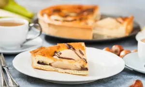Une part de tarte poire chocolat, garnie de fruits et de copeaux de chocolat, est servie avec une tasse de café et des noisettes.