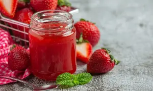 Un pot de confiture de fraise maison, entouré de fruits frais et d'une feuille de menthe. Une cuillère est prête à déguster.