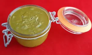 Un pot en verre ouvert révèle une délicieuse pâte à tartiner pistache maison, d'une belle couleur verte, sur un fond rouge vif.