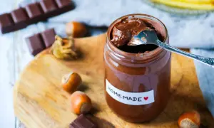 Un pot de pâte à tartiner maison, avec une cuillère, des noisettes et des carrés de chocolat sur une planche en bois.
