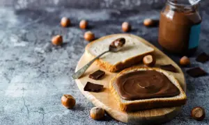 Tranche de pain généreusement tartinée de pâte à tartiner maison 3 ingrédients. Des noisettes et des carrés de chocolat parsèment la table.