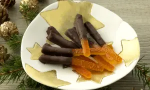Bâtonnets d'orange confite chocolatés sur une assiette étoilée, entourés de pommes de pin dorées et de branches de sapin.