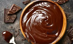 Un bol en bois déborde de chocolat fondu, prêt à travailler. Des éclats de chocolat et une cuillère en argent complètent cette scène gourmande.