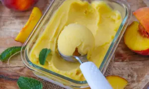 Une boule de glace à la pêche est prélevée d'un bac en verre, entourée de fruits frais et de feuilles de menthe.