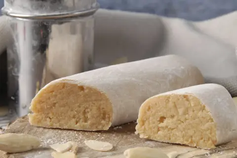 Pâte d'amande prête à être utilisée pour recouvrir un gâteau. Des amandes effilées et un tamis à sucre complètent cette scène gourmande.