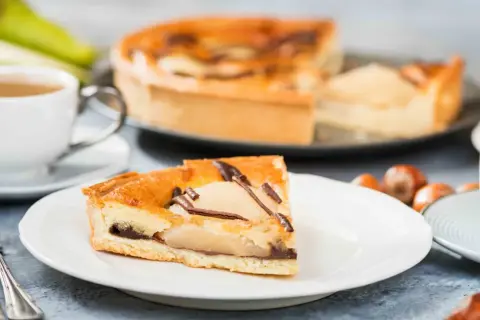 Une part de tarte poire chocolat, garnie de fruits et de copeaux de chocolat, est servie avec une tasse de café et des noisettes.