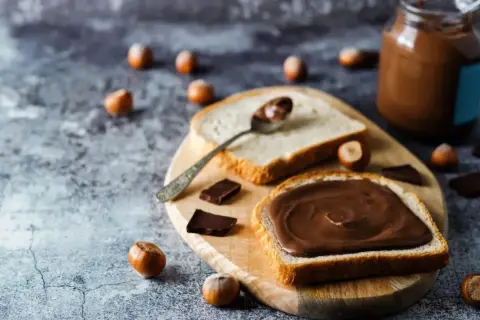 Tranche de pain généreusement tartinée de pâte à tartiner maison 3 ingrédients. Des noisettes et des carrés de chocolat parsèment la table.