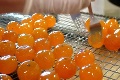Des fruits orangés brillants, prêts à être confits, sont disposés sur une grille. Une fourchette les manipule délicatement.