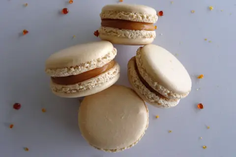 Quatre macarons au caramel beurre salé, parsemés de quelques éclats de caramel, sur fond gris.