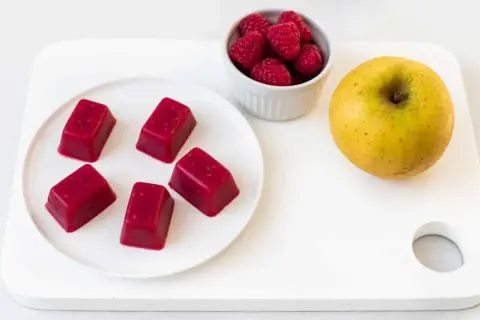 Cinq carrés de pâte de fruit rouge, des framboises et une pomme jaune sur une planche blanche. Une idée de conservation des fruits.
