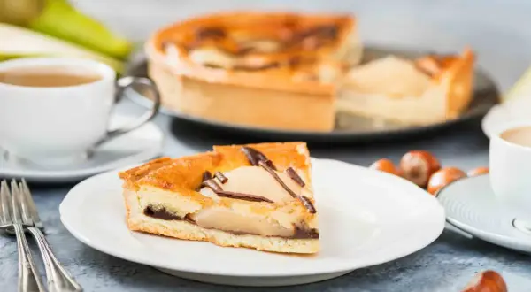 Une part de tarte poire chocolat, garnie de fruits et de copeaux de chocolat, est servie avec une tasse de café et des noisettes.