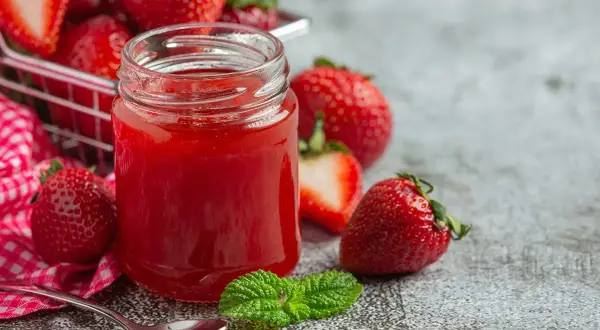 Un pot de confiture de fraise maison, entouré de fruits frais et d'une feuille de menthe. Une cuillère est prête à déguster.