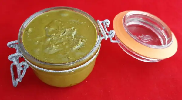 Un pot en verre ouvert révèle une délicieuse pâte à tartiner pistache maison, d'une belle couleur verte, sur un fond rouge vif.