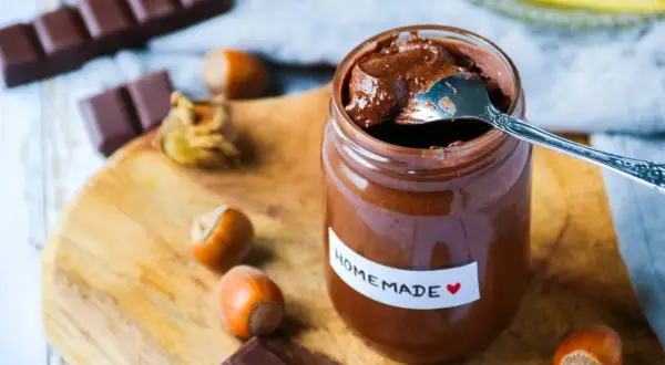Un pot de pâte à tartiner maison, avec une cuillère, des noisettes et des carrés de chocolat sur une planche en bois.