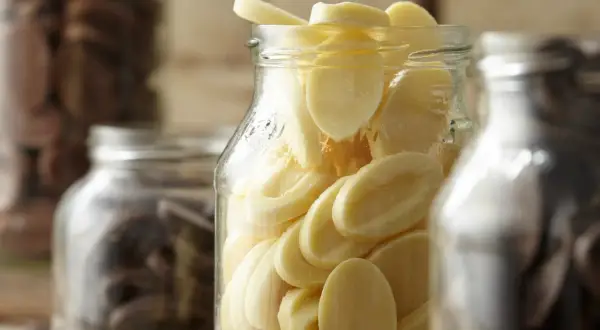 Des éclats de chocolat blanc dans un bocal, prêts pour la conservation chocolat maison. Des fèves de cacao dans d'autres bocaux en arrière-plan.