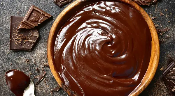 Un bol en bois déborde de chocolat fondu, prêt à travailler. Des éclats de chocolat et une cuillère en argent complètent cette scène gourmande.