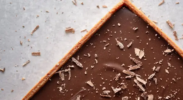 Une délicieuse tarte au chocolat vegan, garnie de copeaux de chocolat, sur un fond clair parsemé de miettes.