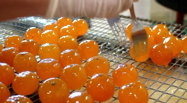 Des fruits orangés brillants, prêts à être confits, sont disposés sur une grille. Une fourchette les manipule délicatement.