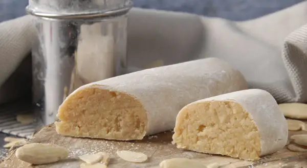 Pâte d'amande, c'est quoi ? Deux rouleaux de massepain, saupoudrés de sucre glace, sur une planche en bois avec des amandes effilées.