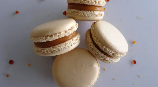 Quatre macarons au caramel beurre salé, parsemés de quelques éclats de caramel, sur fond gris.