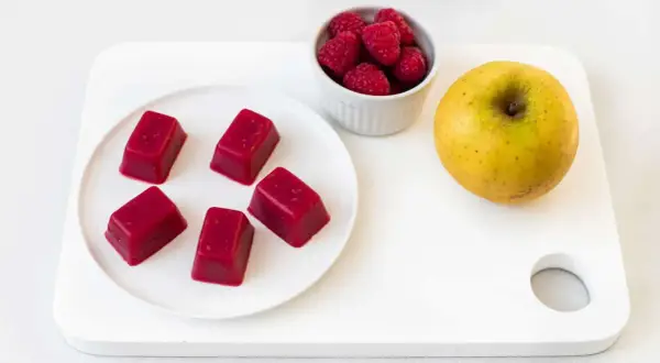 Cinq carrés de pâte de fruit rouge, des framboises et une pomme jaune sur une planche blanche. Une idée de conservation des fruits.