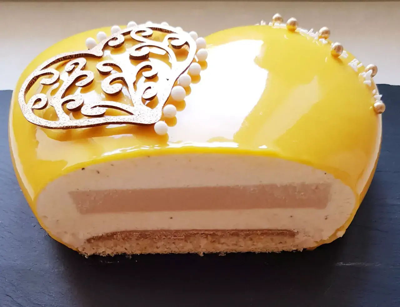 Entremet cœur jaune vif, décoré d'une arabesque dorée et de perles. La coupe révèle des couches onctueuses et un insert caramel beurre salé.