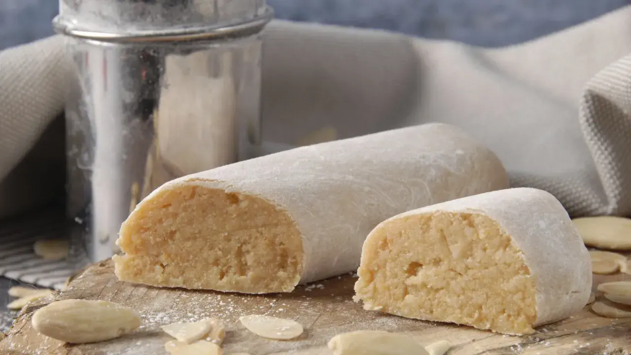Pâte d'amande prête à être utilisée pour recouvrir un gâteau. Des amandes effilées et un tamis à sucre complètent cette scène gourmande.