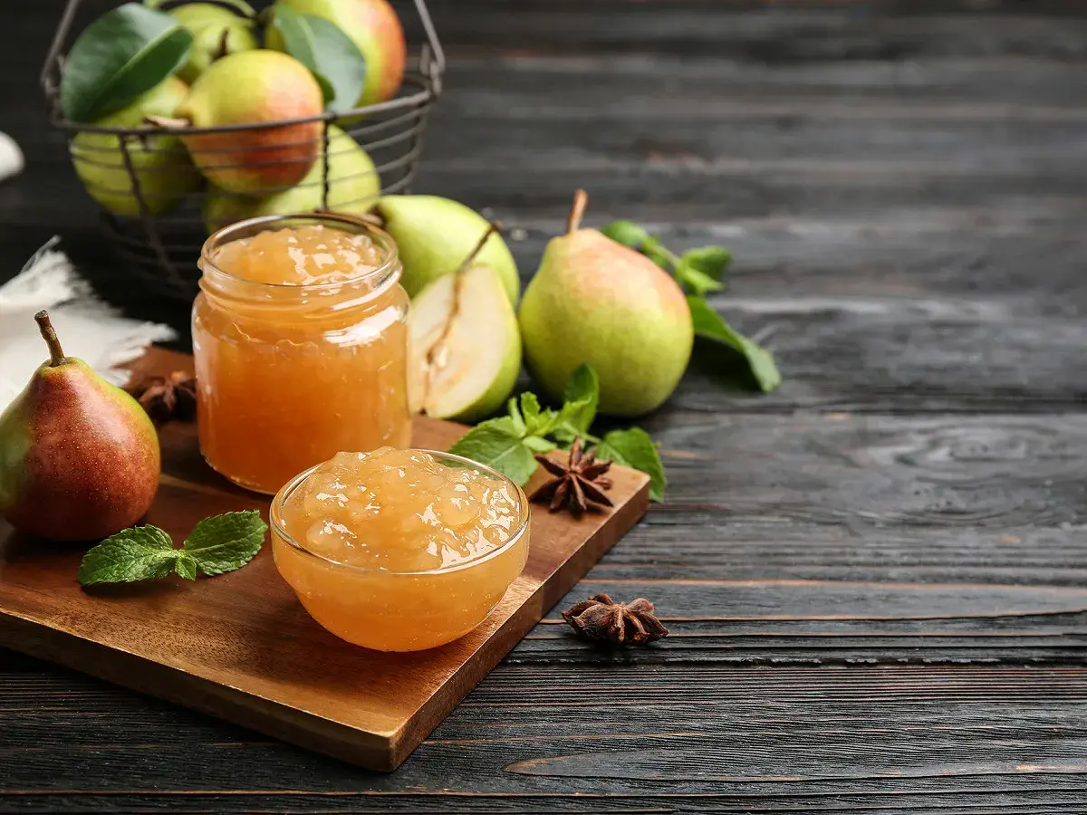 Confiture de poires dans un bocal et un bol, avec des poires fraîches et des feuilles de menthe sur une planche en bois.