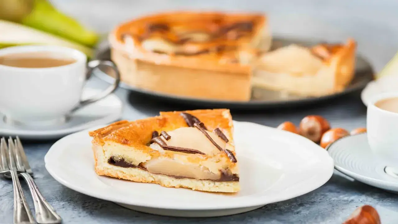 Une part de tarte poire chocolat, garnie de fruits et de copeaux de chocolat, est servie avec une tasse de café et des noisettes.