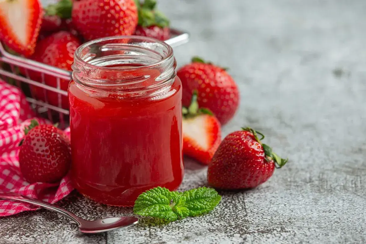 Un pot de confiture de fraise maison, entouré de fruits frais et d'une feuille de menthe. Une cuillère est prête à déguster.