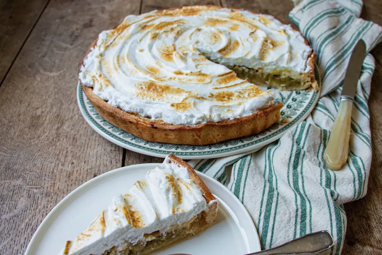 Une part de tarte rhubarbe meringuée, avec sa meringue dorée et sa garniture acidulée, sur une assiette blanche.