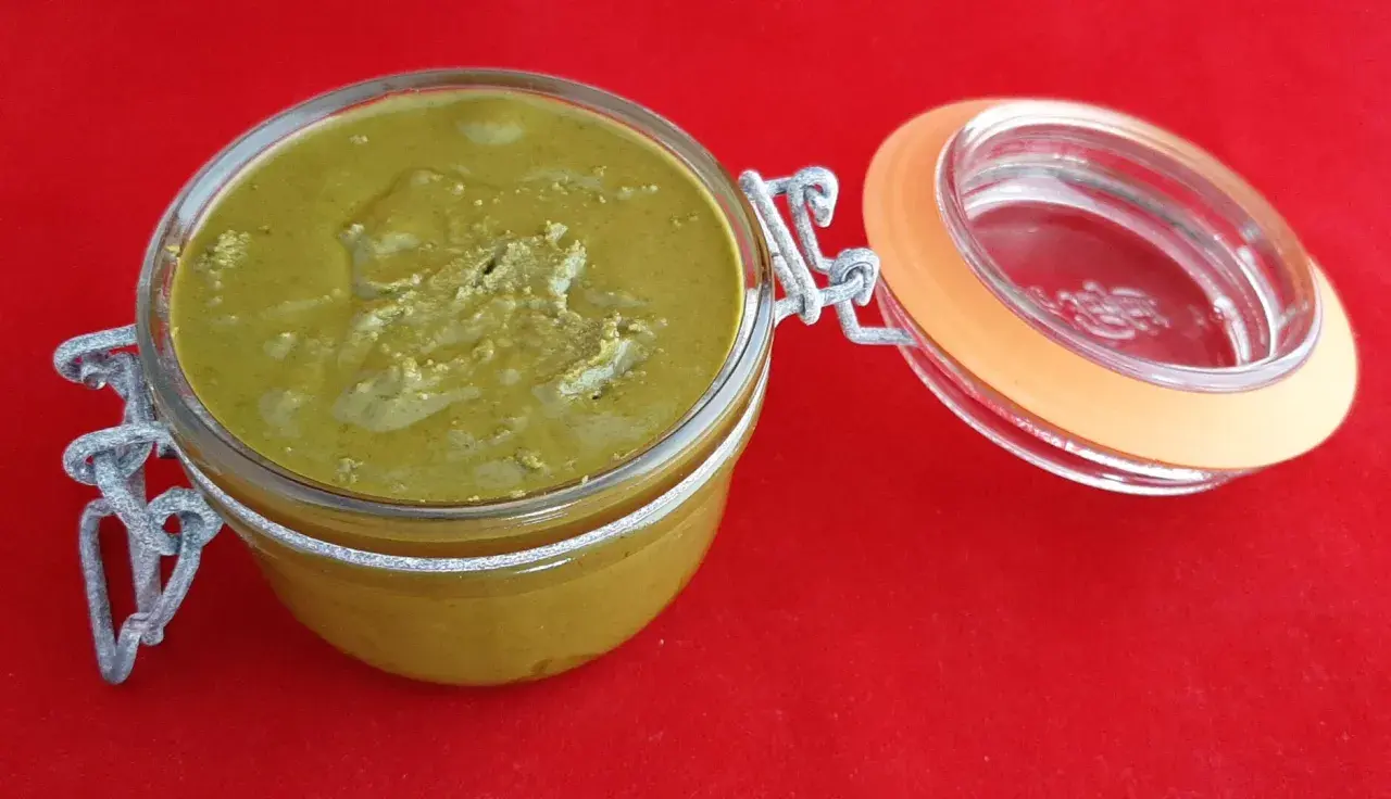 Un pot en verre ouvert révèle une délicieuse pâte à tartiner pistache maison, d'une belle couleur verte, sur un fond rouge vif.