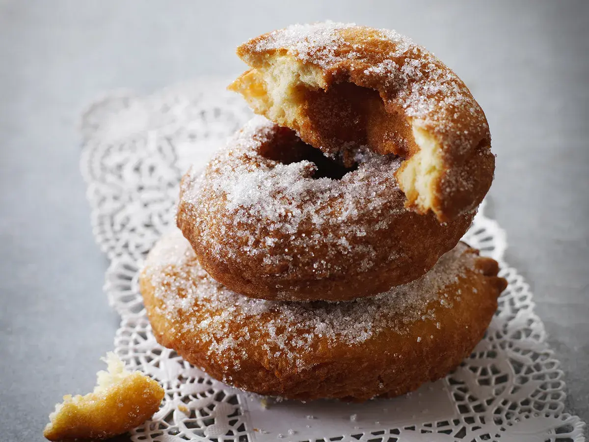 Un beignet au sucre, moelleux et doré, est empilé sur un autre, saupoudré de cristaux de sucre. Un morceau croqué révèle sa texture aérienne.