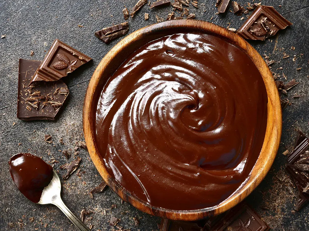 Un bol en bois déborde de chocolat fondu, prêt à travailler. Des éclats de chocolat et une cuillère en argent complètent cette scène gourmande.