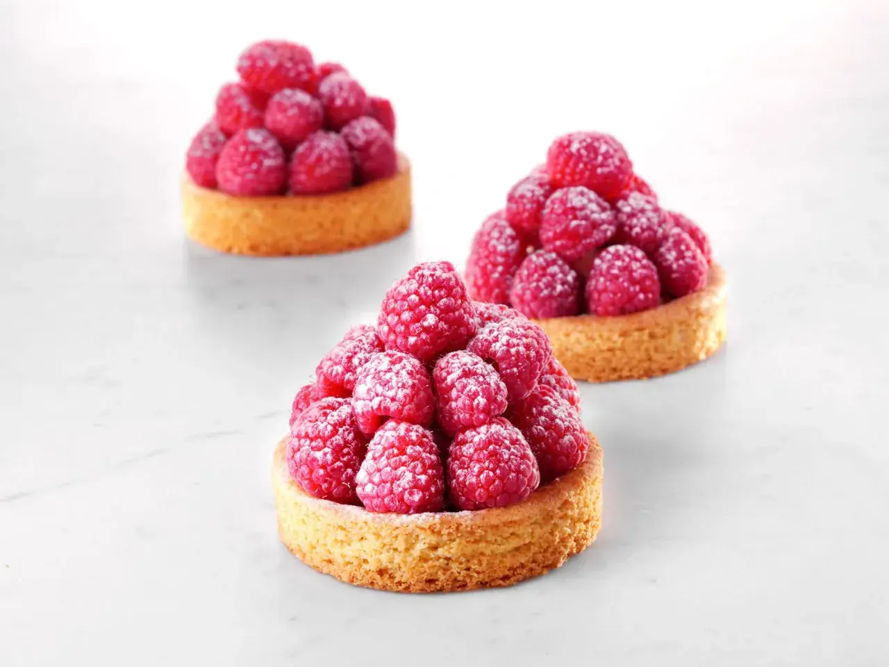 Trois tartelettes sablé breton garnies de framboises fraîches saupoudrées de sucre glace. Une invitation à la gourmandise.