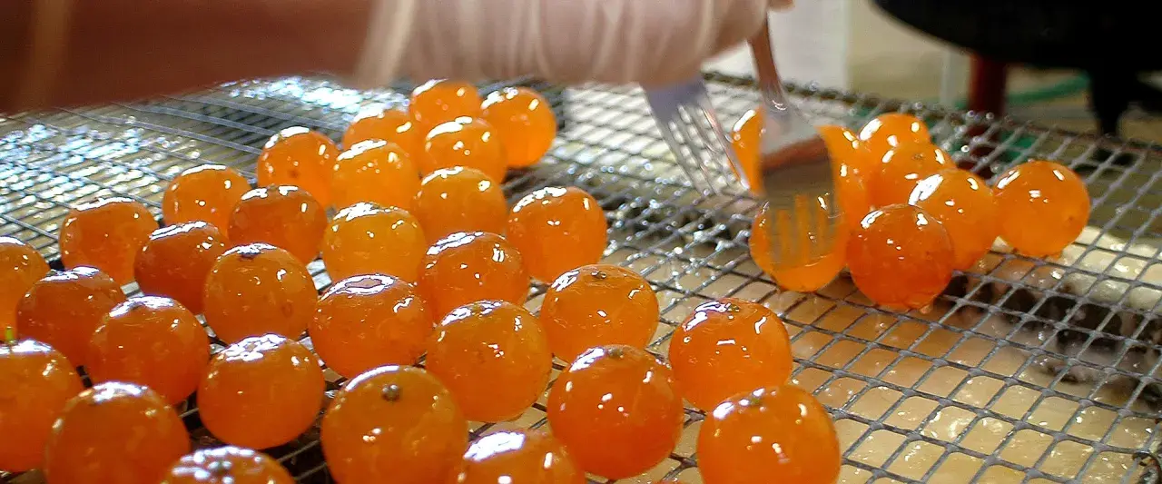 Des fruits orangés brillants, prêts à être confits, sont disposés sur une grille. Une fourchette les manipule délicatement.