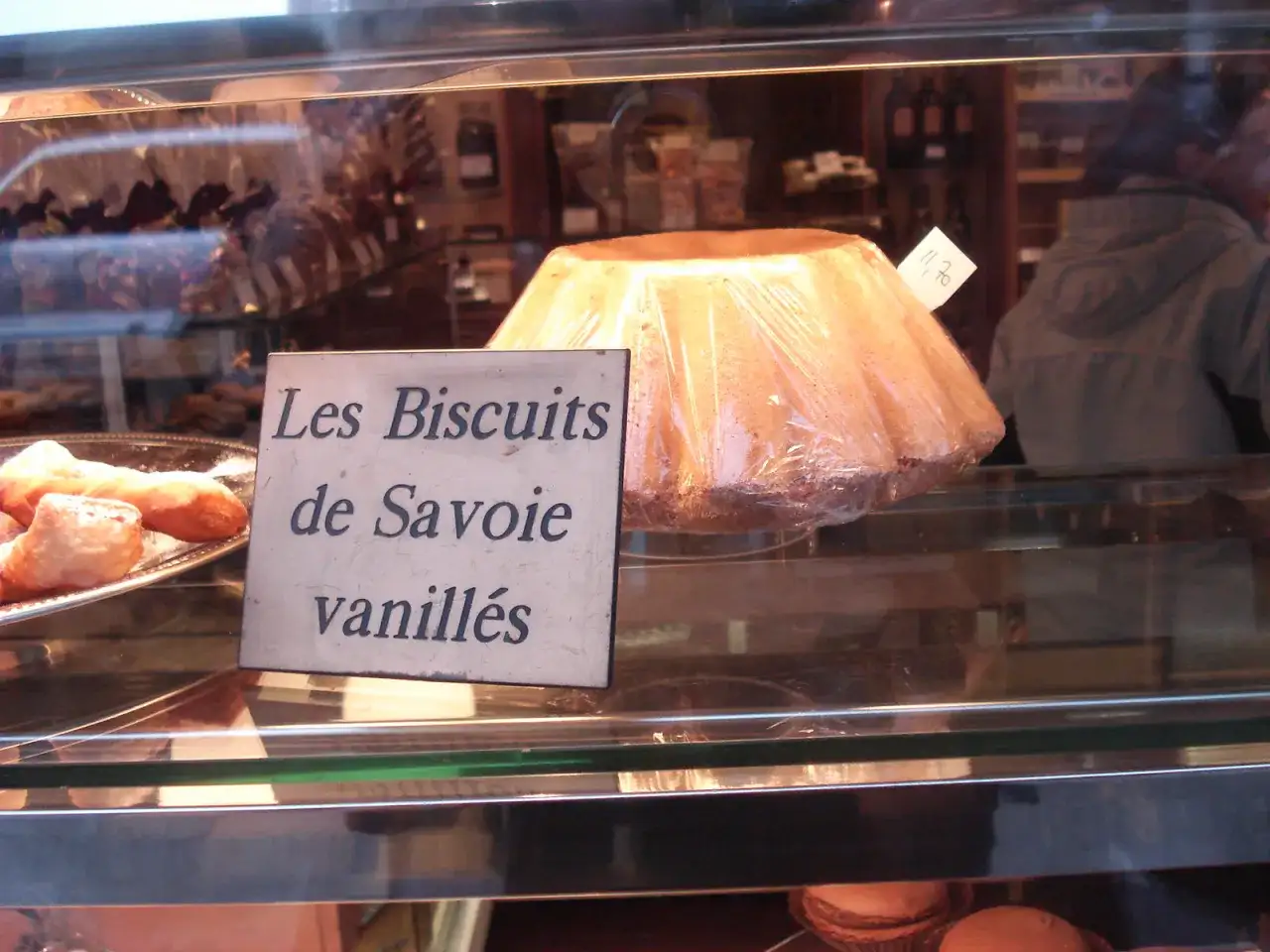 Un grand biscuit de Savoie vanillé, emballé sous plastique, est présenté dans une vitrine de boulangerie.