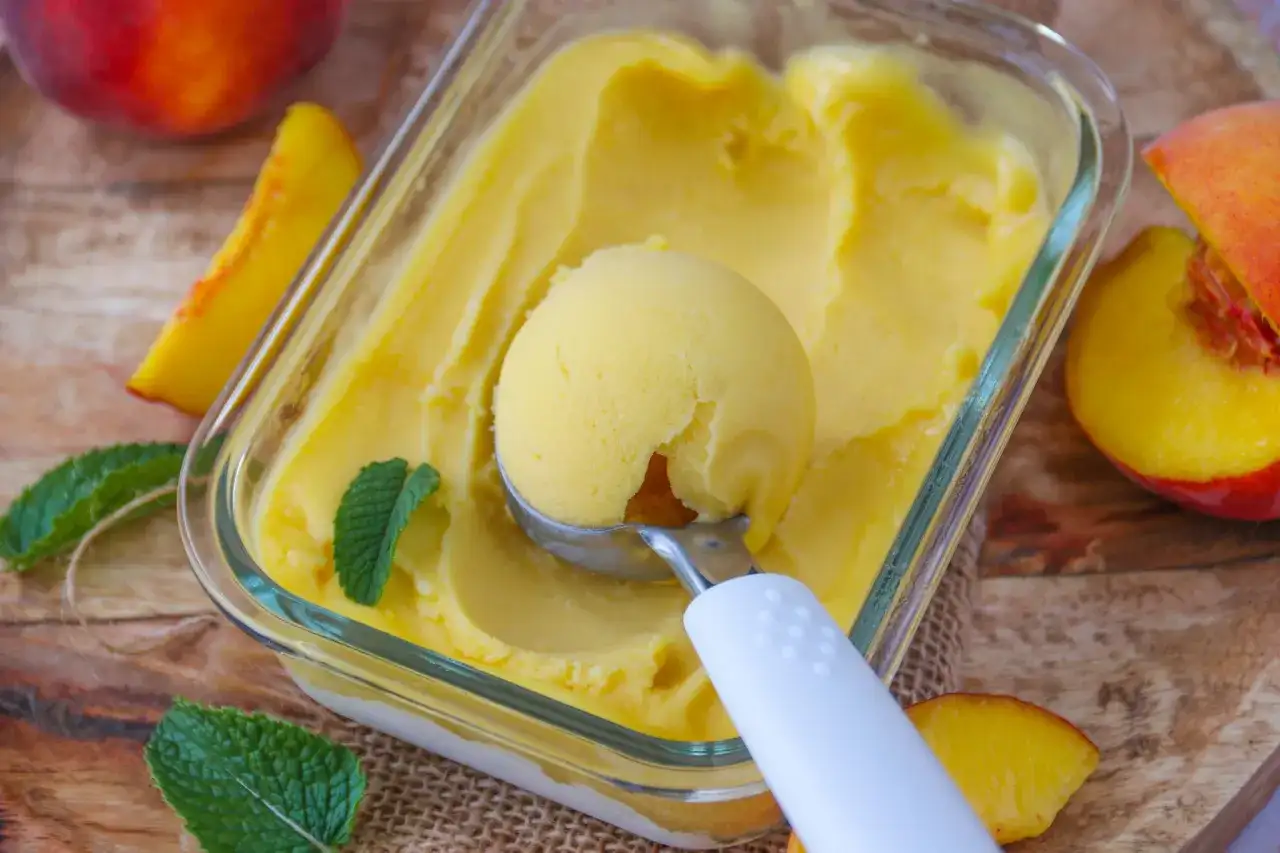 Une boule de glace à la pêche est prélevée d'un bac en verre, entourée de fruits frais et de feuilles de menthe.