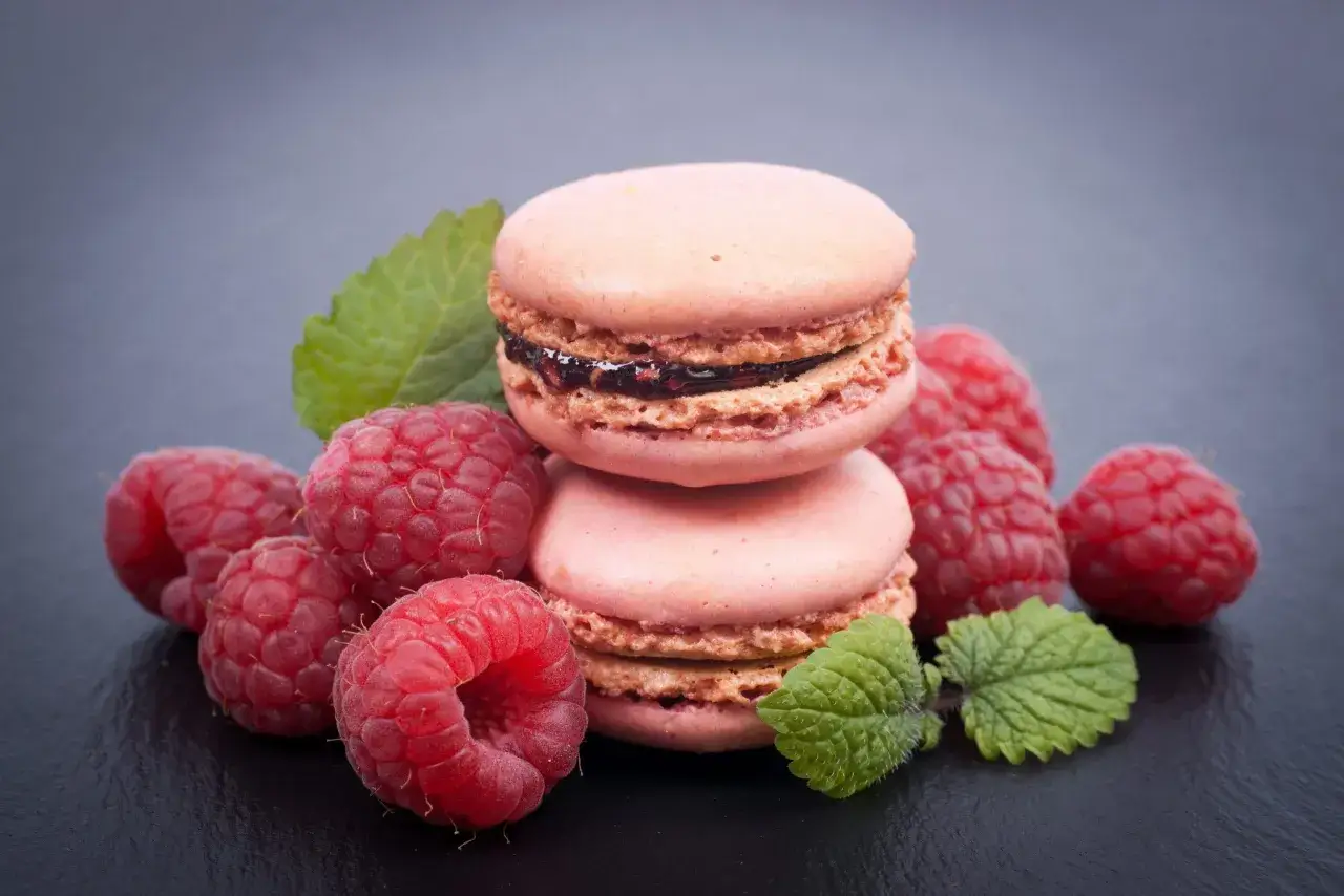 Macarons roses garnis d'une ganache framboise et d'une touche de gélatine, entourés de framboises fraîches et de feuilles de menthe.