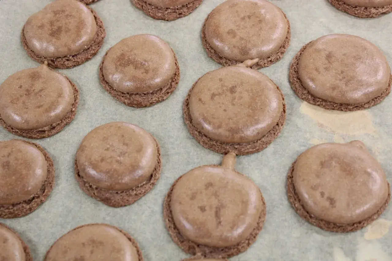 Des macarons bruns, certains avec des pointes, pos&eacute;s sur une feuille de cuisson. Une texture qui peut expliquer pourquoi les macarons craquent.