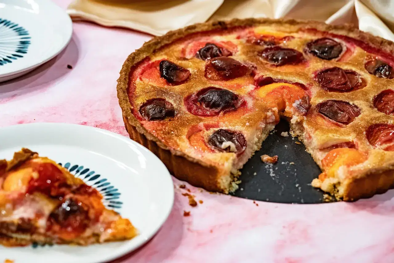 Une délicieuse tarte aux prunes, dorée et garnie de fruits juteux, est prête à être dégustée. Une part a déjà été servie sur une assiette.