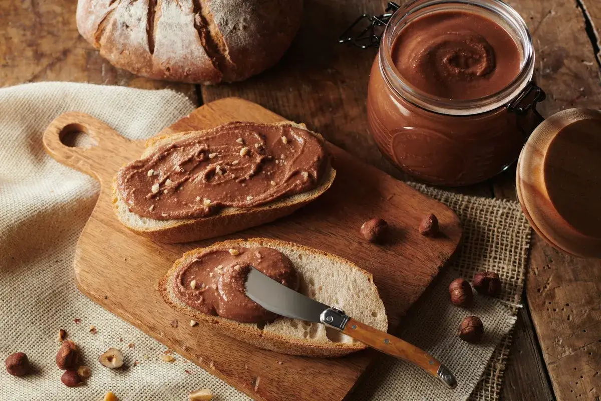 Tranches de pain de campagne tartinées de pâte à tartiner maison, avec un pot de cette gourmandise et des noisettes.