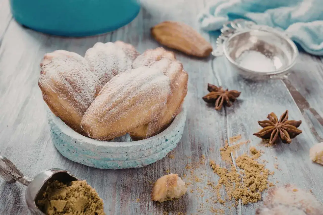 Des madeleines moelleuses saupoudrées de sucre glace dans un bol bleu, avec des anis étoilés et une cuillère de poudre sur une table en bois.