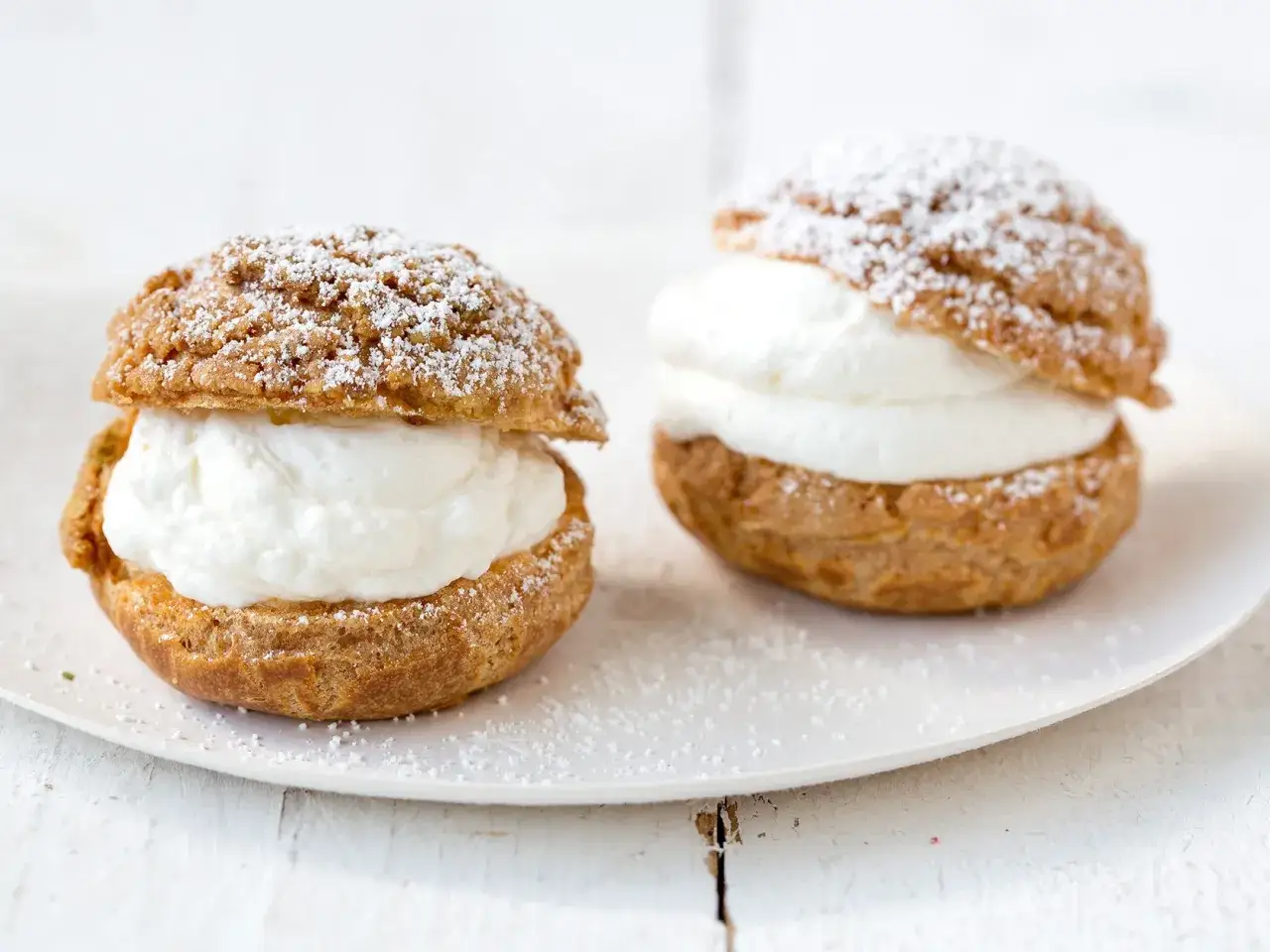 Deux choux à la crème généreusement garnis de crème fouettée, saupoudrés de sucre glace, sur une assiette blanche.