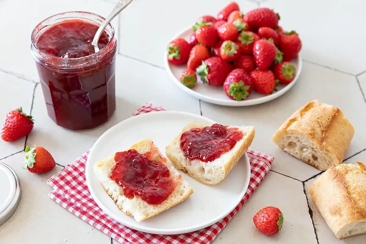 Un pot de confiture de fraise ouvert, des tartines de pain garnies de confiture, des fraises fraîches et des morceaux de baguette. Un délice !