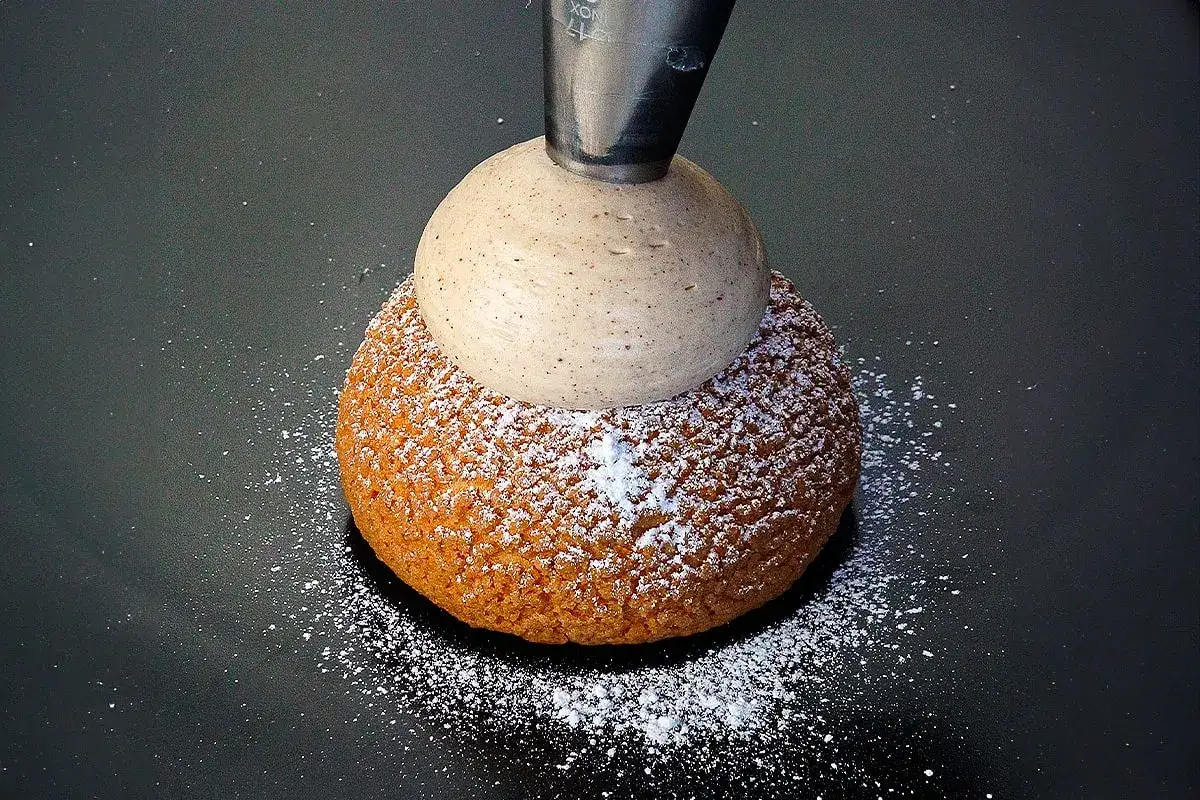 Une poche &agrave; douille d&eacute;pose une g&eacute;n&eacute;reuse boule de ganache mont&eacute;e chocolat sur un chou saupoudr&eacute; de sucre glace.