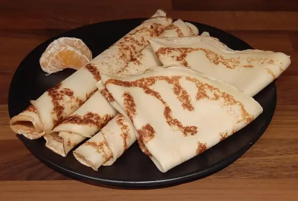 Crêpes dorées, roulées et pliées, sur une assiette noire, accompagnées de quelques quartiers d'orange. Un délice de crêpe sans oeuf.