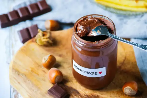 Un pot de pâte à tartiner maison, avec une cuillère, des noisettes et des carrés de chocolat sur une planche en bois.