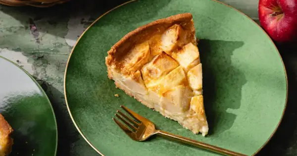 Une part de tarte normande aux pommes, dorée et appétissante, sur une assiette verte avec une fourchette dorée.