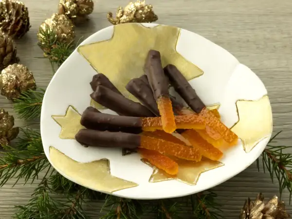 Bâtonnets d'orange confite chocolatés sur une assiette étoilée, entourés de pommes de pin dorées et de branches de sapin.